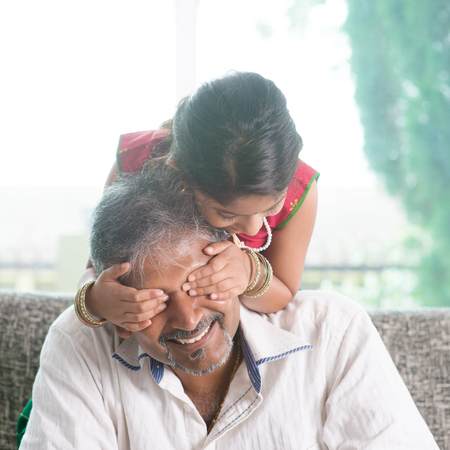 Happy Indian family at home. Asian girl surprising her father by covering daddy eyes. Parent and child indoor lifestyle.の写真素材