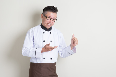 Portrait of 50s mature Asian male chef in uniform showing something and giving thumb up, standing on plain background with shadow, copy space.の写真素材