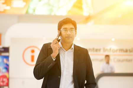 Asian Indian businessman walking by information counter, talking on phone, golden sunset background.の写真素材