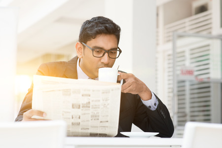 Young Asian Indian businessman sipping coffee and reading newspaper at cafe. India male business man, modern office building with beautiful golden sunlight as background.の写真素材