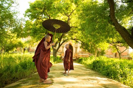 Two little Buddhist novice monks walking outdoors under shade of green tree, outside monastery, Myanmar.の写真素材