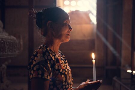 Old wrinkled traditional Asian burmese woman praying with candle light inside a temple, low light,...