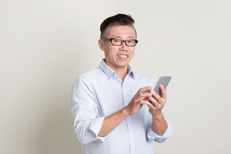 Portrait of single mature 50s Asian man in casual business texting using smartphone and smiling, standing over plain background with shadow. Chinese senior male people.の写真素材