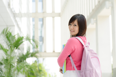 Young Asian teen student standing outside school campus building, holding file folder and smiling.の写真素材