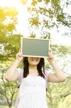 Young Asian college girl student standing on campus lawn, hands holding a blank chalkboard covering face and smiling.の写真素材