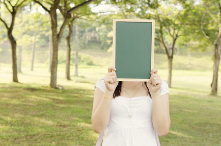 Young Asian college girl student standing on campus lawn, hands holding a blank chalkboard and covered face.の写真素材
