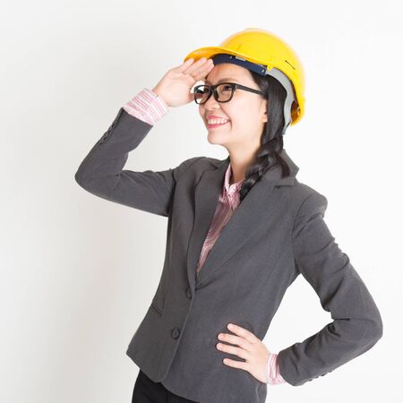 Portrait of Asian female engineer with hard hat hand shielding and looking away, standing on plain background.の写真素材
