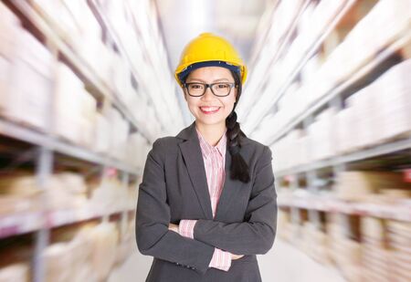Young Asian female store manager with hard hat arms crossed standing in storehouse. Shelves with goods at background.の写真素材
