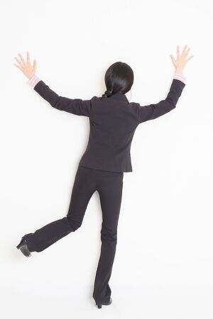 Full body back view of young Asian businesswoman in formalwear banging and leaning on wall, standing on plain background.の写真素材