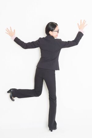 Full length back view of young Asian businesswoman in formalwear hands and body leaning on wall, standing on plain background.の写真素材