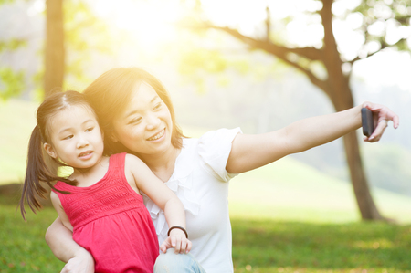 Lifestyle portrait of Asian mother and daughter taking selfie in happiness in the park, family outdoor fun, morning with sun flare.の写真素材