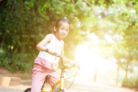 Portrait of active Asian child riding bicycle outdoors. Little girl having fun at nature park. Morning sun flare background.の写真素材