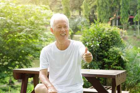 Portrait of cheerful gray hair Asian old man giving thumbs up, sitting at outdoor park in morning.の写真素材