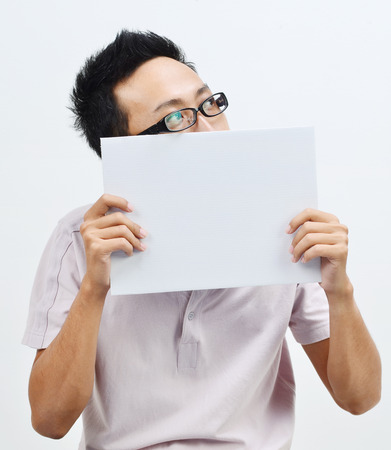 Portrait of Asian man hand holding white blank paper card covering his mouth and looking at side, standing isolated on plain background.の写真素材