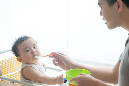 Happy Asian family at home. Father feeding solid food to his 9 months old baby in the kitchen,...
