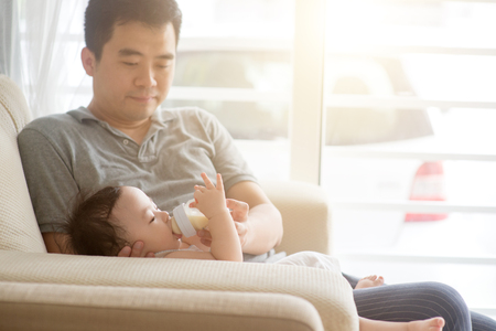 Happy Asian family at home. Father bottle feed formula milk to 9 months old baby boy, living lifestyle indoors. の写真素材