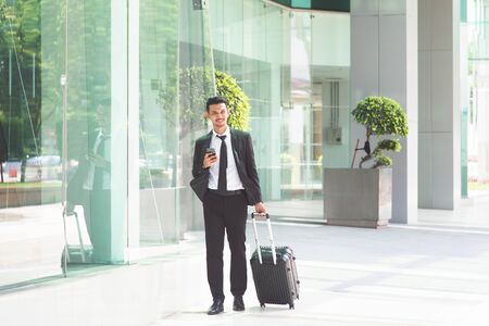 Busy Asian businessman with luggage walking to airport, checking schedule from smartphone.の写真素材