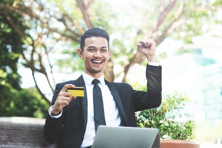 Happy Asian business man cheering, hand holding credit card sitting with laptop outdoor.の写真素材