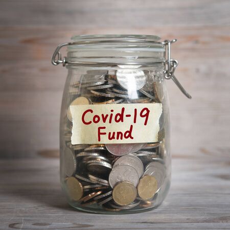 Coins in glass money jar with covid19 fund label. Vintage wooden background with dramatic light.の写真素材