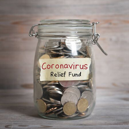 Coins in glass money jar with Coronavirus relief fund label, financial concept. Vintage wooden background with dramatic light.の写真素材