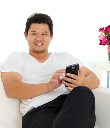 Asian man sitting on sofa and using handphoneの写真素材