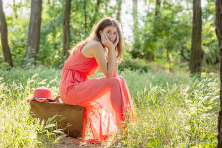 Young woman in red dress sitting on old brown suitcase and looking into camera in the woods with backlightの写真素材