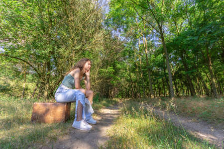 Young woman sitting on an old brown suitcase in a forest dirt roadの写真素材