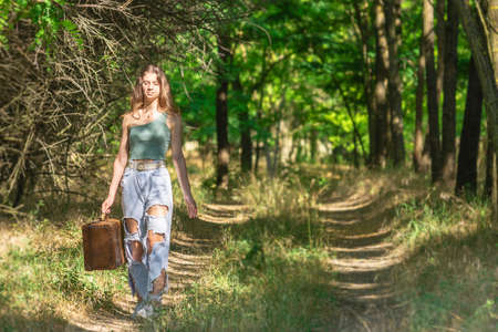Young woman walking in forest dirt road with old brown suitcaseの写真素材