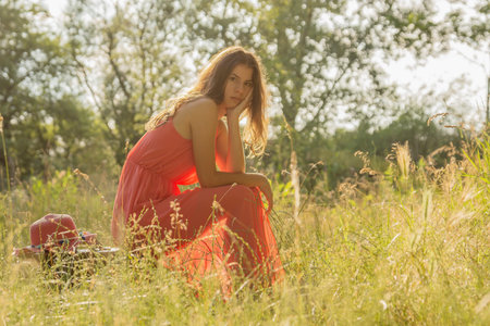 Young woman in red dress sitting on suitcase and looking into camera in the woods with backlightの写真素材