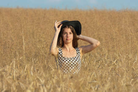 Young woman with black hat standing and looking into camera half-covered in wheat field on a sunny dayの写真素材