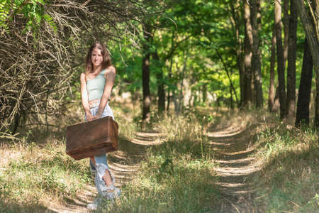 Young woman standing in forest dirt road with old brown suitcaseの写真素材
