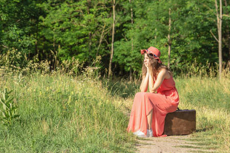 Young woman in red dress with hat sitting on old brown suitcase in forest dirt roadの写真素材