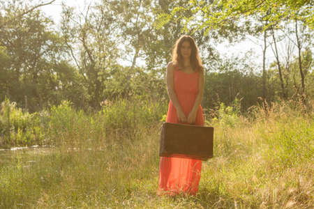 Backlight young woman in red dress standing with old brown suitcase and looking into cameraの写真素材