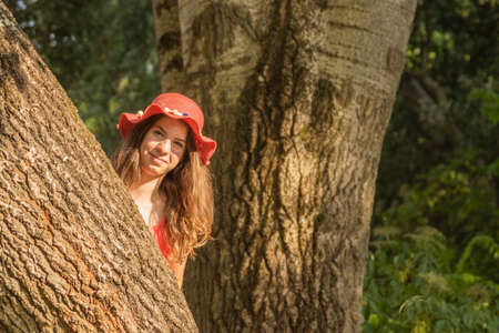 Young woman in red dress and hat looking into camera behind a tree trunkの写真素材