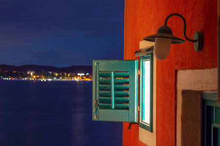 Window of a shuttered house in Rovinj old town at night and behind it a sea bay with a small lights, Croatiaの写真素材