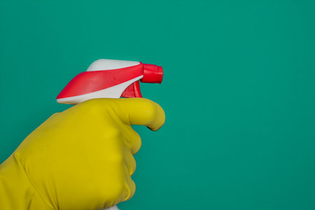 Hand in yellow rubber glove holding plastic spray bottle with cleaning detergent on blue background