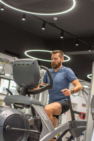An active man with beard training in gym, riding a stationary exercise bike. Sport and quarantine.