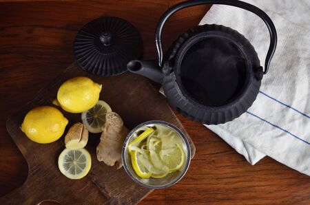 Overhead view of a black iron tea pot with hot water, fresh ginger and lemon on a cutting board, a white kitchen cloth and a white Chinese tea set with two cups on a dark wooden table.の写真素材