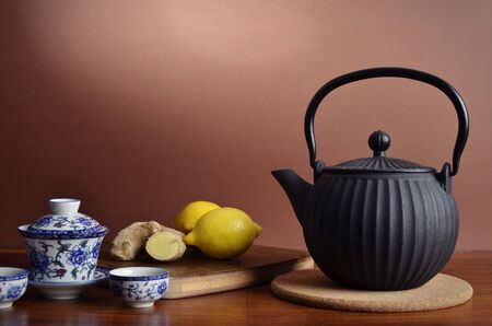 A black iron tea pot, fresh ginger and lemon on a cutting board and a white Chinese tea set with two cups on a dark wooden table, with brown backgroundの写真素材