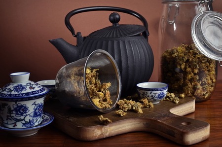 Side view of a black iron tea pot, accompanied by a white chinese tea set and a tea strainer with tea in it, on a wood cutting board, with a glass container in the background with chrysanthemum flower tea.の写真素材