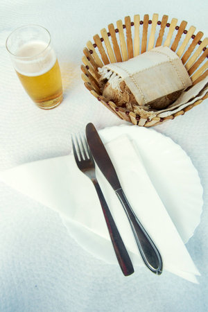 Fork and knife over white plate and napkin with beer and bread basketの写真素材