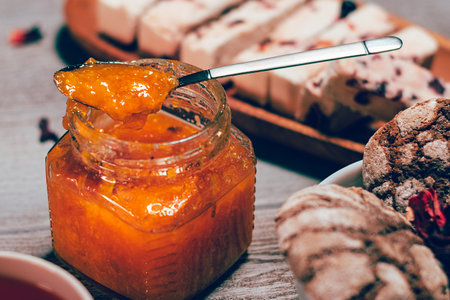 Orange jam jar on wooden table close up. Blurred chocolate biscuit and marshmellow on background