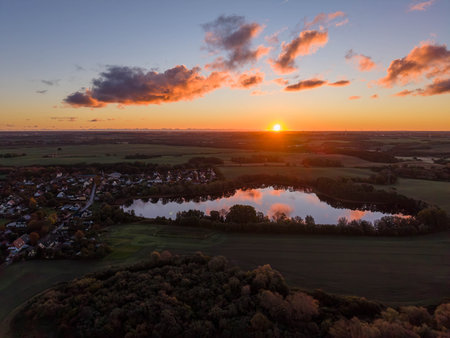 still lake mirrors the glowing sunset sky, surrounded by autumn trees, open fields, and a quiet village nestled in the serene rural landscapeの写真素材