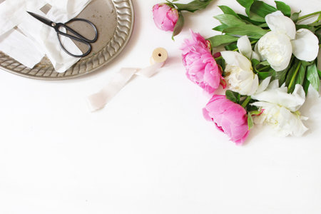 Styled stock photo. Feminine wedding or birthday table composition with floral bouquet. White and pink peonies flowers, old vintage scissors, silver tray and silk ribbons. White background.の写真素材