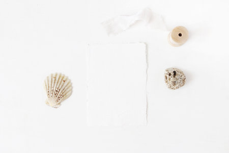 Summer marine, wedding stationery, desktop mock-up scene. Blank greeting cotton paper card, seashells, pebble stone and wooden spool of silk ribbon. White table background. Flat lay, top view.の写真素材