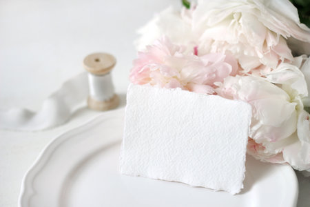 Bright wedding or birthday stationery mockup scene with a handmade paper greeting card, porcelain plate, spool of silk ribbon and pink peony flowers a white table background. Feminine styled stock photography.の写真素材