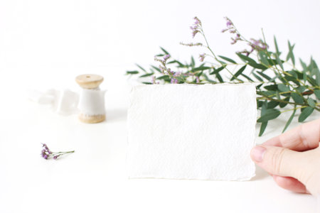 Bright feminine wedding stationery mockup scene. Closeup of woman hand holding handmade paper place card. Spool of silk ribbon, eucalyptus leaves and violet limonium flowers on white table background.の写真素材