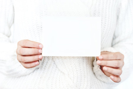 Closeup of womans hands in white knitted sweater holding blank paper card. Greeting card mock-up. Chritsmas winter design. Feminine styled stock photo.の写真素材