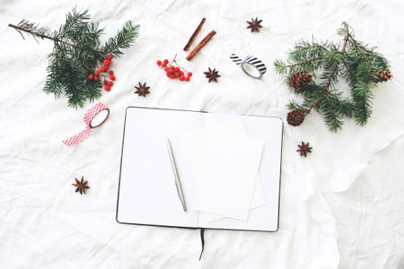 Christmas composition. Blank note pad, greeting cards mock-up scene. Christmas tree branches, red rowan berries, pine cones, washi tapes and cinnamon. White linen tablecloth background. Flat lay, top.の写真素材