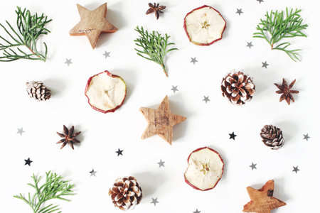 Christmas festive styled composition. Decorative pattern. Pine cones, dried apple fruit, cypress branches, anise, confetti and wooden stars isolated on white wooden background. Flat lay, top viewの写真素材
